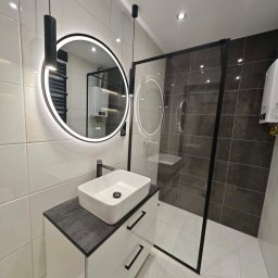 Firma Produkcyjno-Handlowo-Usługowa ROMEX Alicja Niezgoda - Nowoczesna łazienka z białą umywalką nablatową, szafką z szufladami, okrągłym lustrem LED i kabiną prysznicową z czarnymi profilami. Minimalistyczny design.
