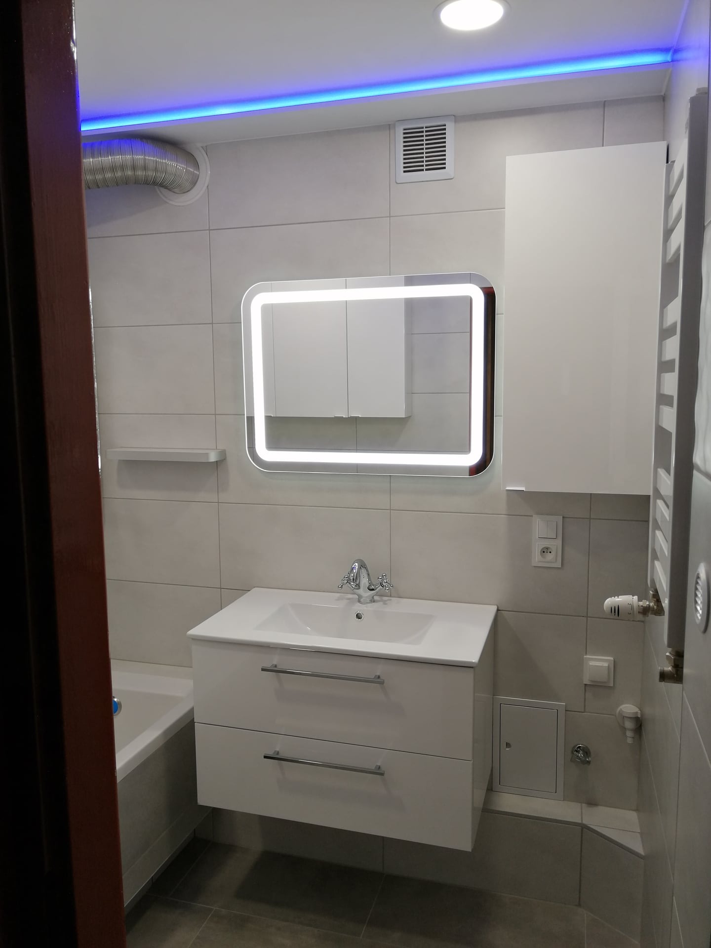 Nowoczesna łazienka w jasnych odcieniach szarości i bieli. Umywalka z szafką, lustro z podświetleniem LED, grzejnik drabinkowy i niebieskie oświetlenie sufitowe.