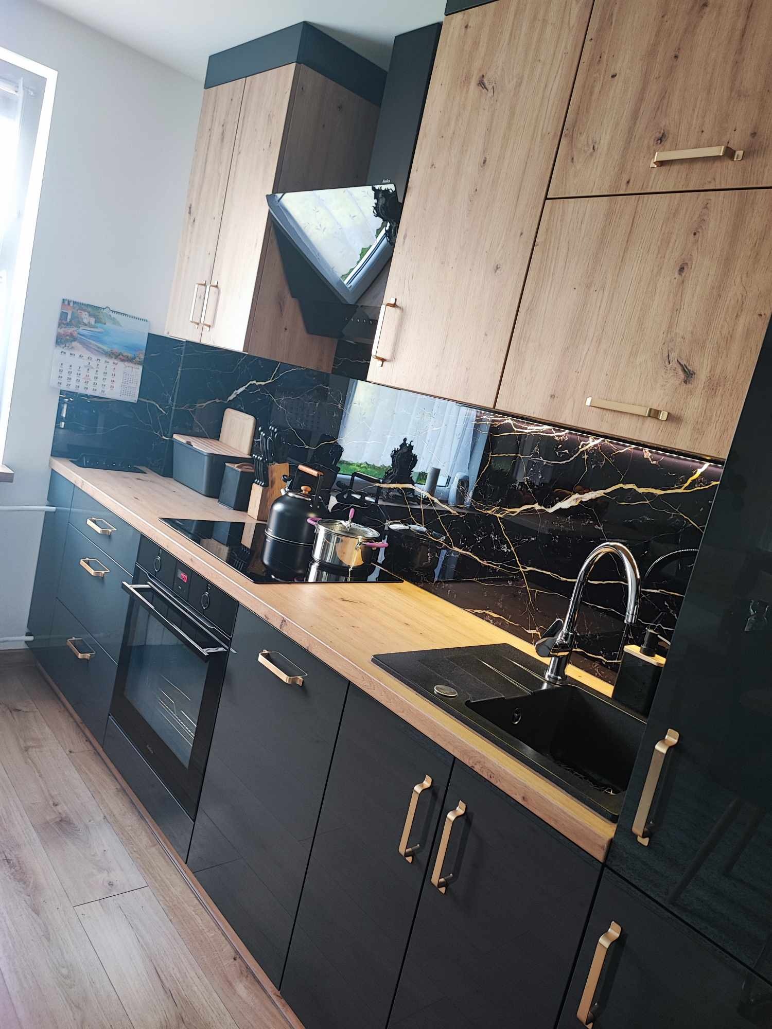 Nowoczesna kuchnia z czarnymi szafkami, drewnianymi blatami i marmurowym backsplash'em z złotymi żyłami. Widoczny czajnik na płycie indukcyjnej, zlew i bateria.