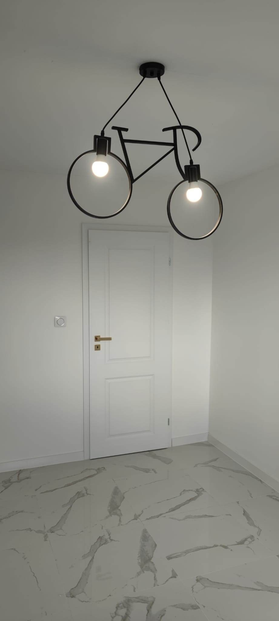 Nowoczesna lampa sufitowa w kształcie roweru z okrągłymi reflektorami, oświetlająca białe drzwi i marmurową podłogę w minimalistycznym wnętrzu.