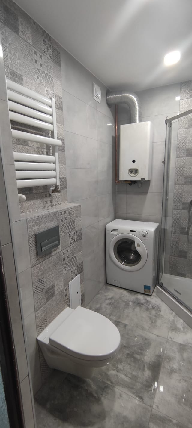 Szara łazienka po remoncie: podwieszana toaleta, grzejnik drabinkowy, pralka i kabina prysznicowa. Płytki w odcieniach szarości z dekoracyjnym wzorem.