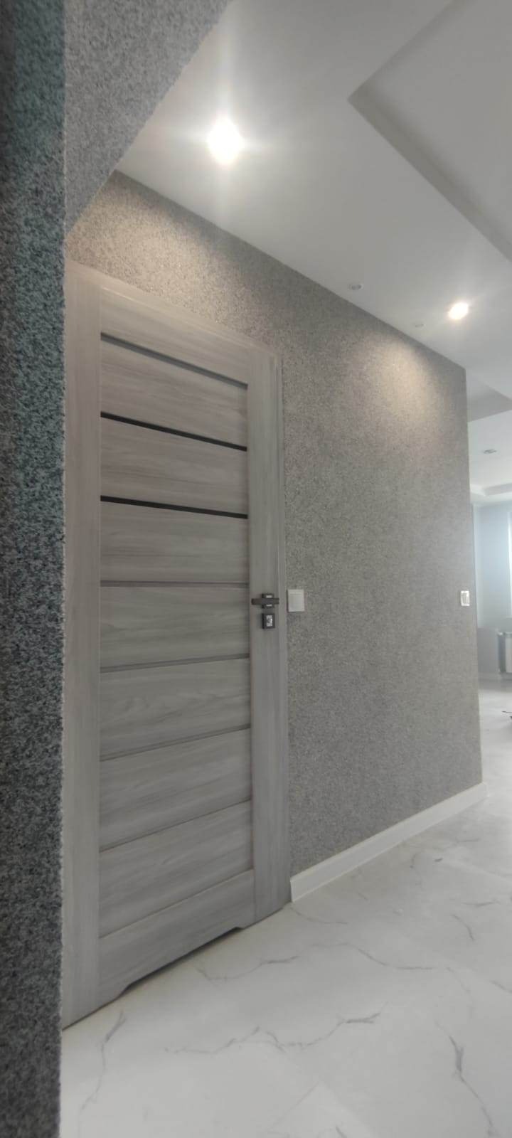 Jasnoszare drzwi z poziomymi wstawkami w wykończonym wnętrzu. Ściany pokryte dekoracyjnym tynkiem w odcieniach szarości. Biała, marmurowa podłoga i oświetlenie sufitowe.