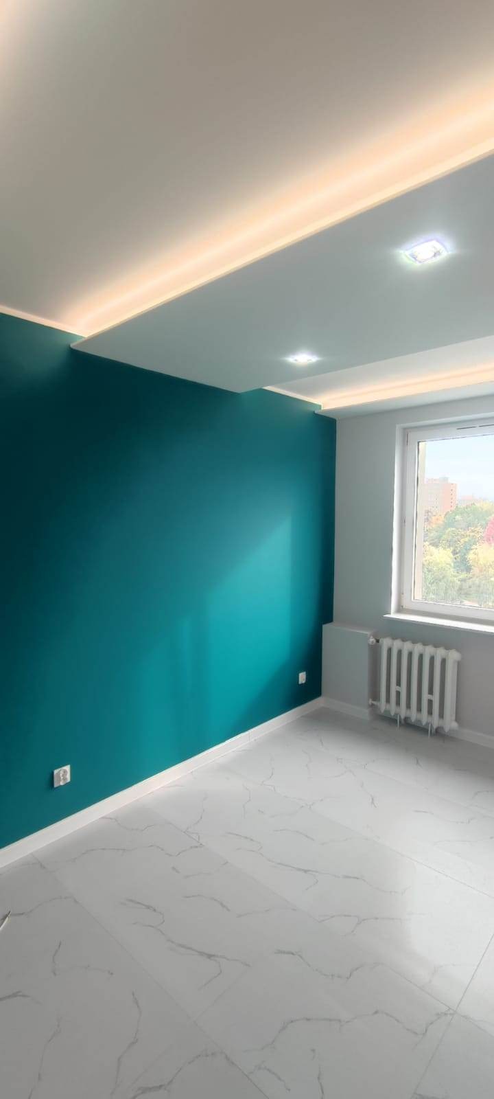 Pomalowane na turkusowo ściany i biała marmurowa podłoga w pustym pokoju z oknem. Sufit podwieszany z oświetleniem LED. Grzejnik w kolorze szarym.