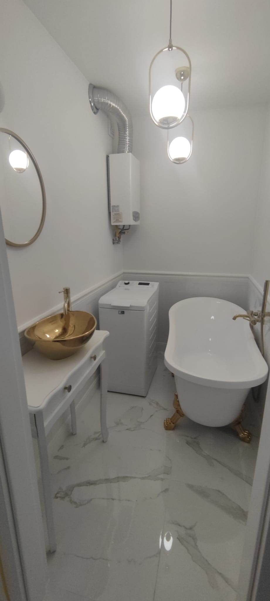 Biała łazienka z wolnostojącą wanną na złotych nóżkach, złotą umywalką na białej konsoli i białą pralką. Oświetlenie stanowią nowoczesne lampy wiszące.