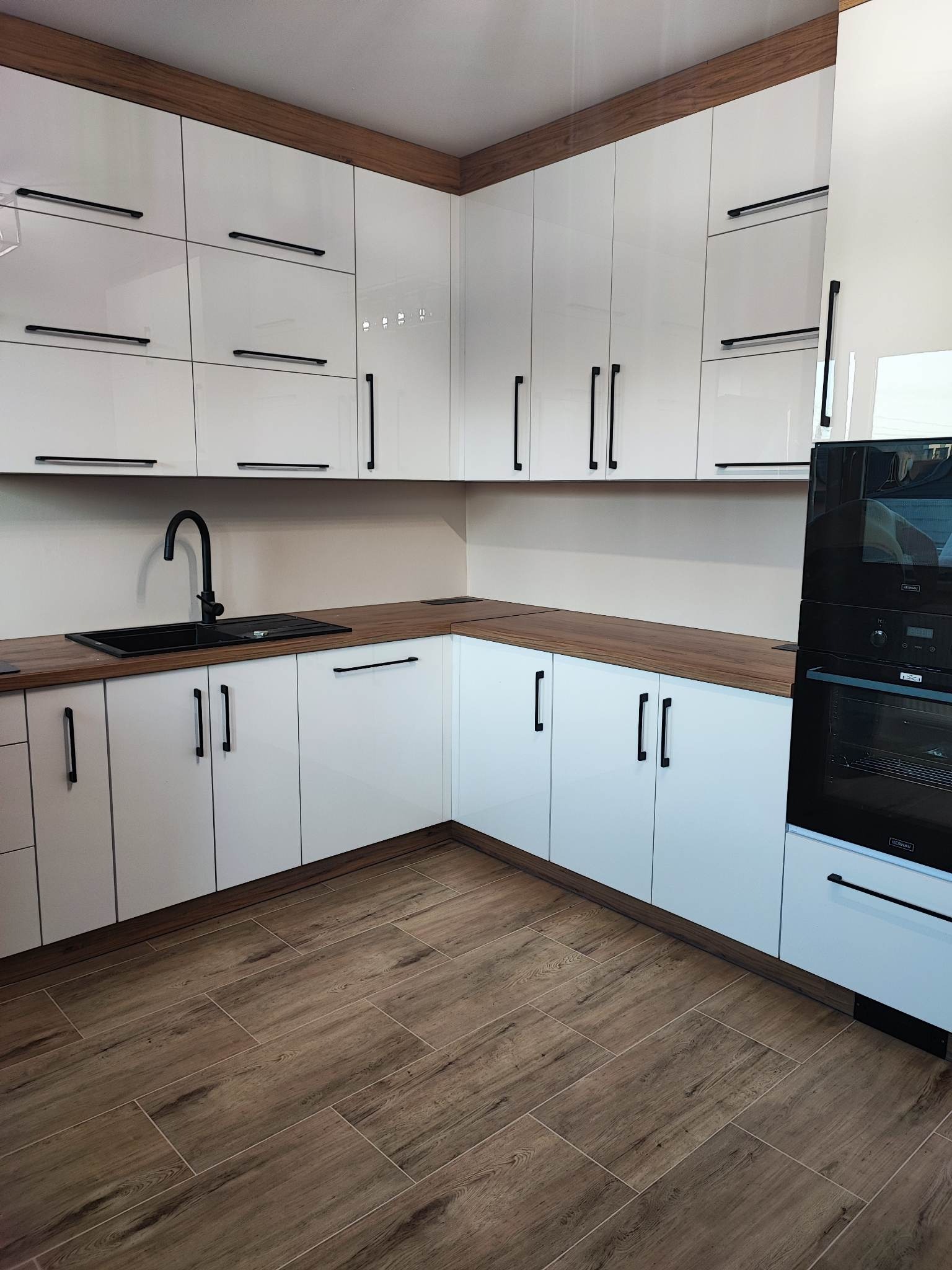 Biała kuchnia na wymiar z drewnianymi blatami i czarnymi uchwytami. Uwagę przykuwa czarny zlew i bateria. Podłoga wyłożona panelami imitującymi drewno.