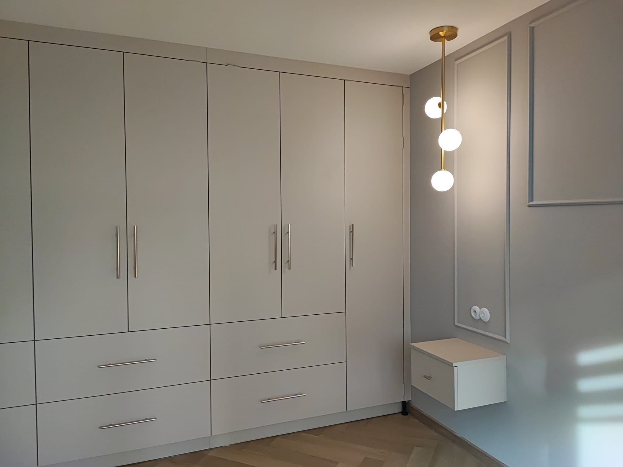 Jasnoszara szafa na wymiar z szufladami i złotymi uchwytami, uzupełniona o minimalistyczną lampę wiszącą i szafkę nocną, na tle ściany z dekoracyjnymi listwami.