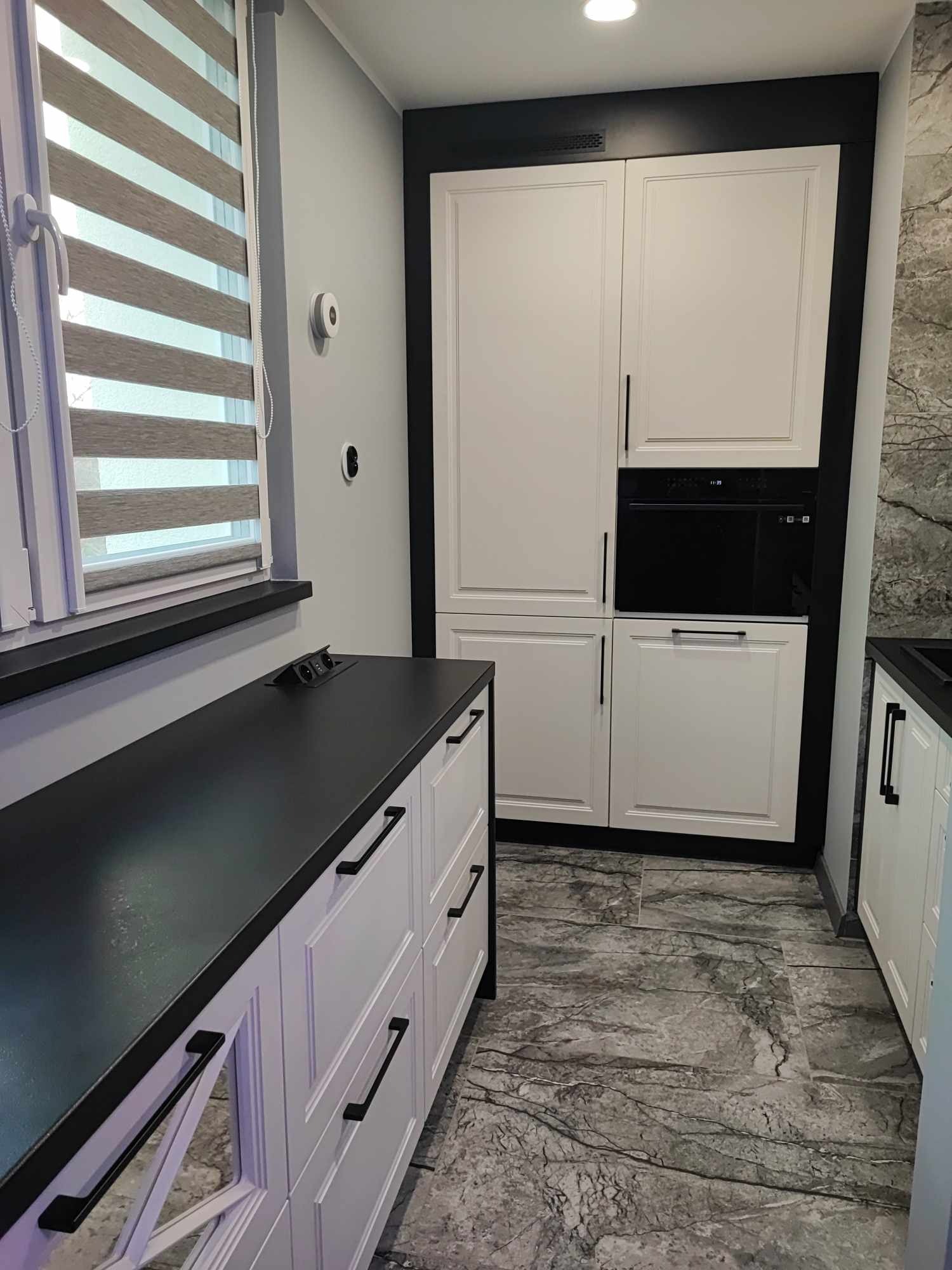 Nowoczesna kuchnia na wymiar z białymi frontami, czarnym blatem i zabudowaną piekarnikiem. Widoczne żaluzje okienne i marmurowe płytki podłogowe.