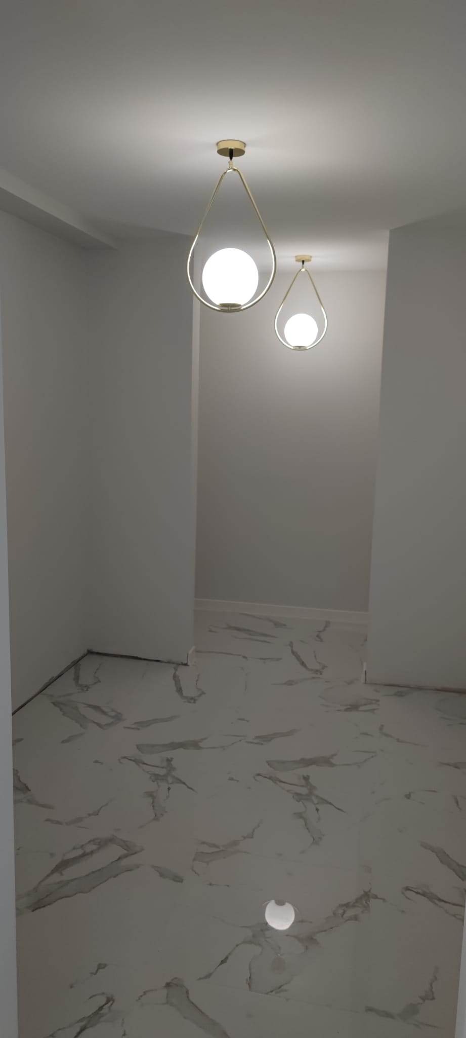 Nowoczesne oświetlenie: Dwie lampy sufitowe w kształcie łez z kulistymi kloszami i złotymi oprawami w minimalistycznym, jasnym wnętrzu z marmurową podłogą.