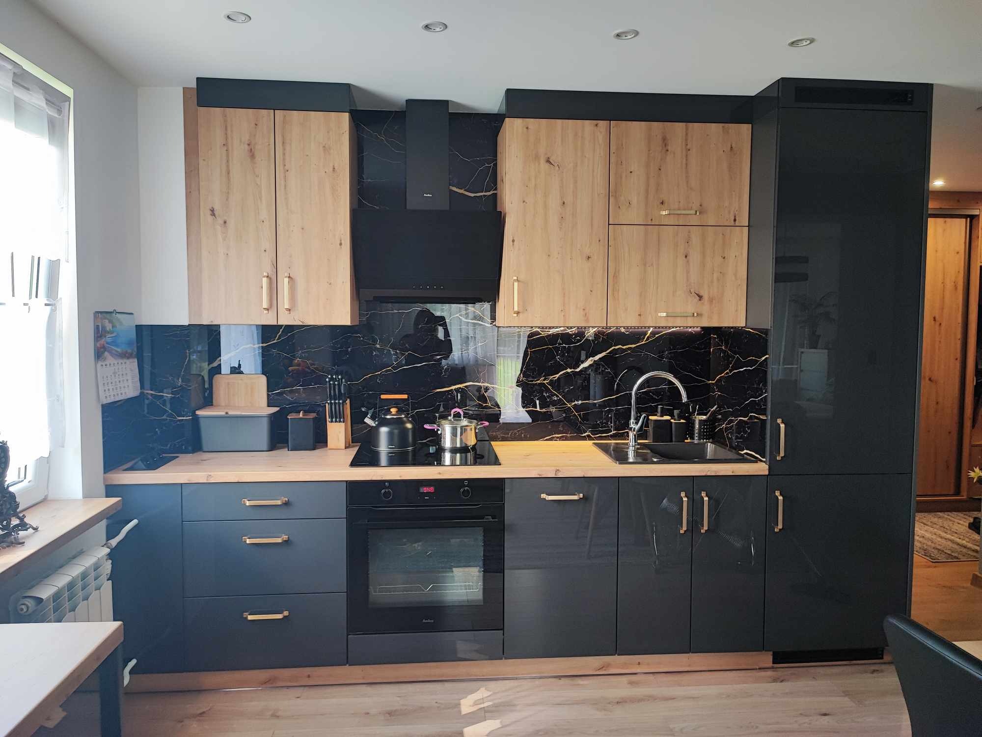 Nowoczesna kuchnia z czarnymi lakierowanymi frontami i drewnianymi akcentami. Blat drewniany, czarny panel z marmurowym wzorem, złote uchwyty. Widoczny zlew i płyta.