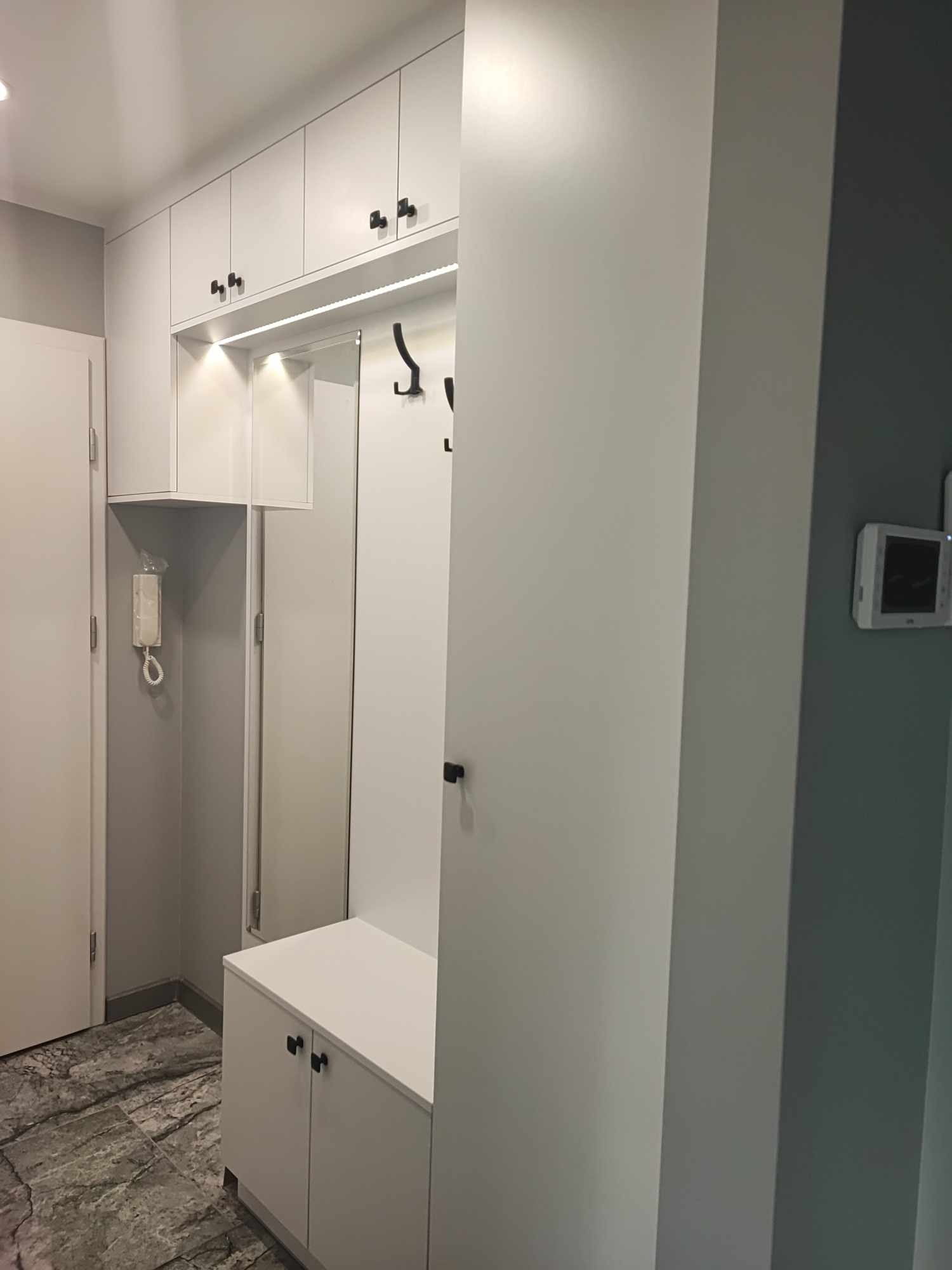 Biała szafa wnękowa z siedziskiem i wieszakami w przedpokoju. Szafki górne z oświetleniem LED. Ciemne uchwyty szafek. Na ścianie domofon.