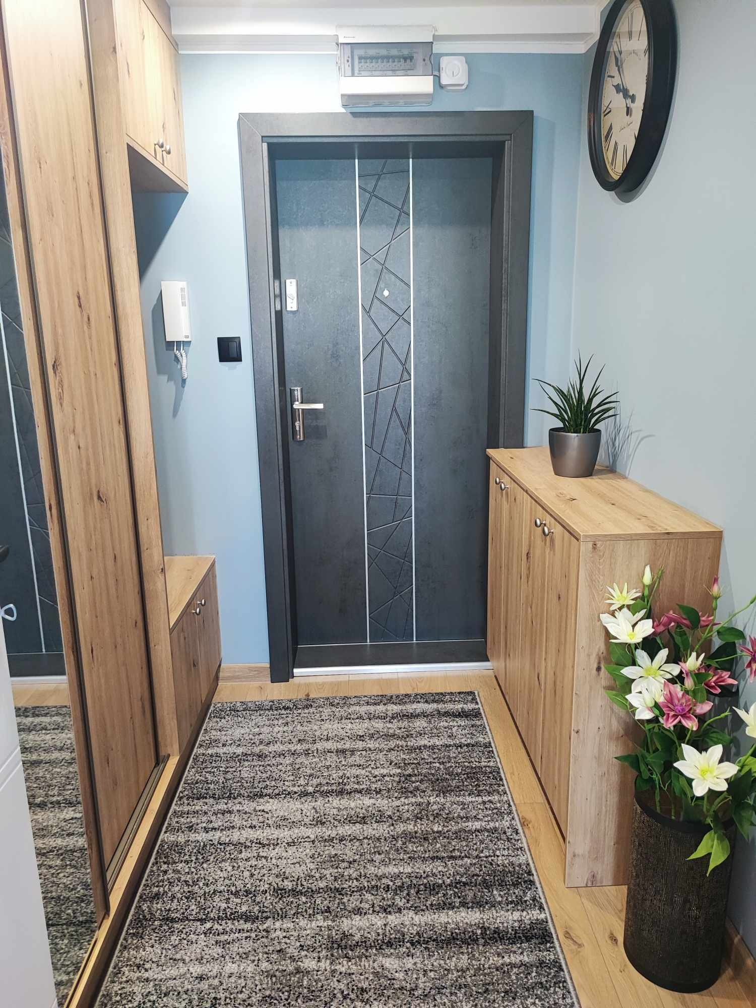 Nowoczesny przedpokój z szafą z lustrem, szafką na buty i szarymi drzwiami z geometrycznym wzorem. Całość dopełnia dywanik i dekoracje roślinne.