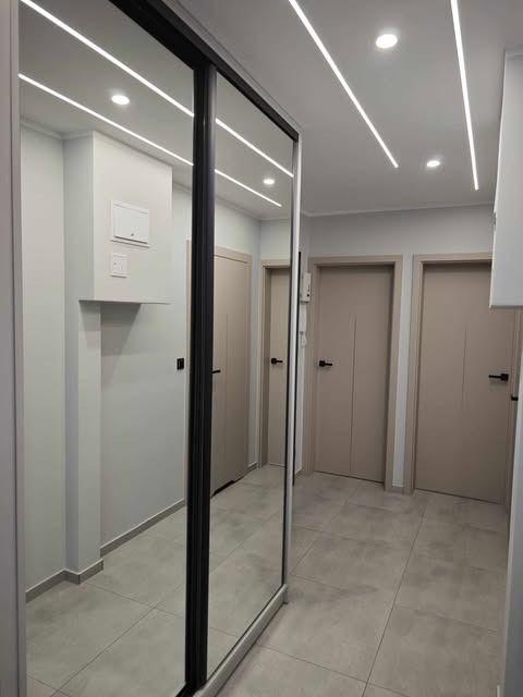 Nowoczesna szafa przesuwna z lustrzanymi drzwiami w przedpokoju. Minimalistyczny design, szara kolorystyka, oświetlenie LED na suficie. Widok na drzwi wejściowe.