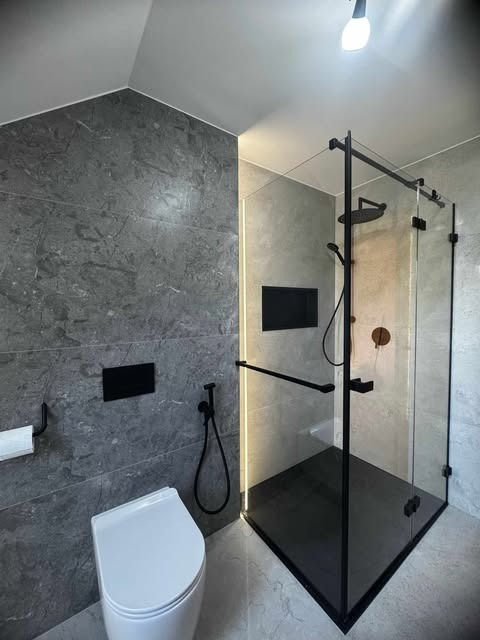 Nowoczesna łazienka z kabiną prysznicową typu walk-in z czarnymi profilami, szarą marmurową okładziną ścian i podwieszaną toaletą. Minimalistyczny design, oświetlenie LED.