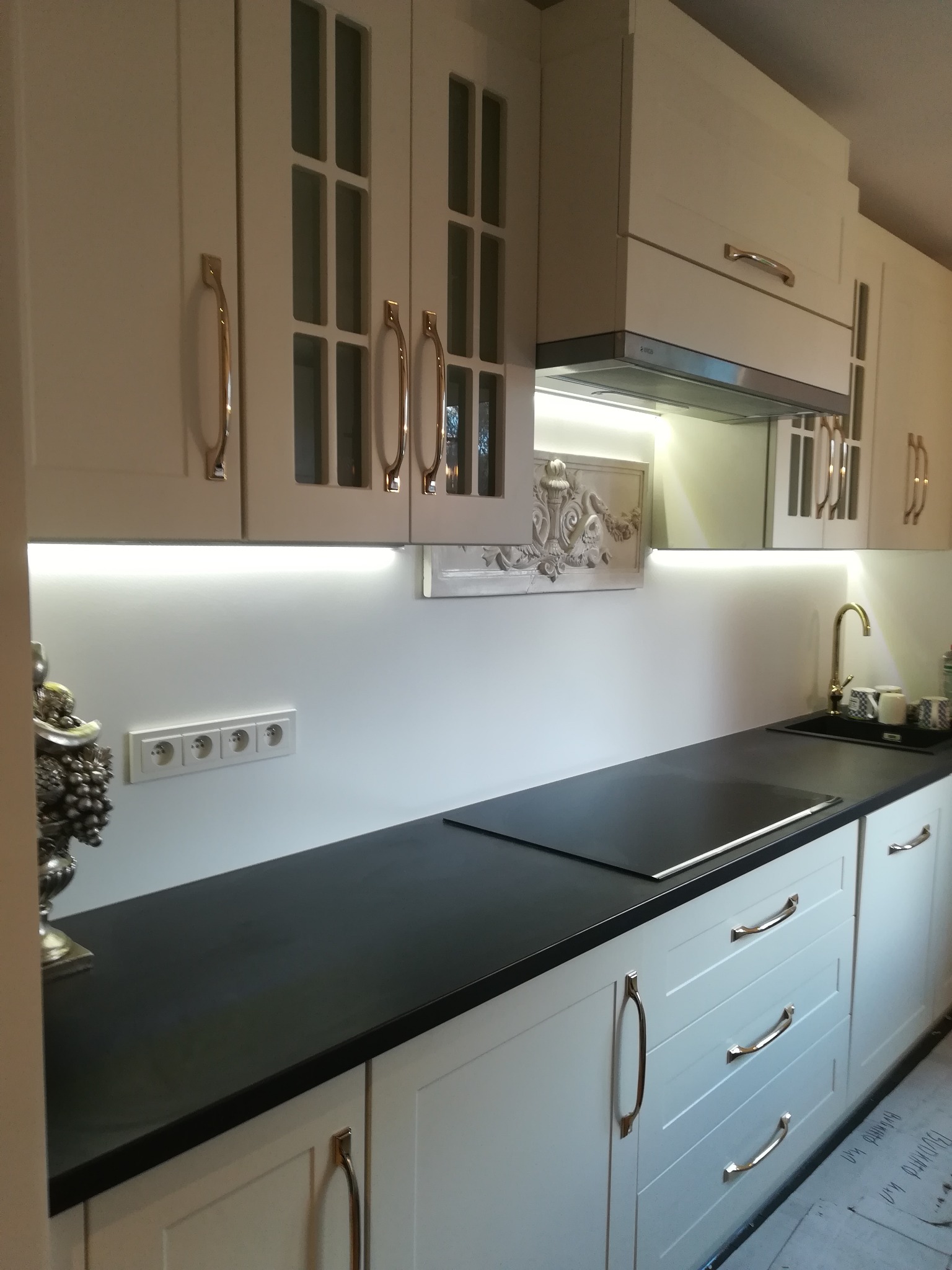 Jasna kuchnia z frezowanymi frontami, czarnym blatem i złotymi uchwytami. Podświetlenie LED pod szafkami górnymi. Dekoracyjny panel nad blatem. Nowoczesny design.