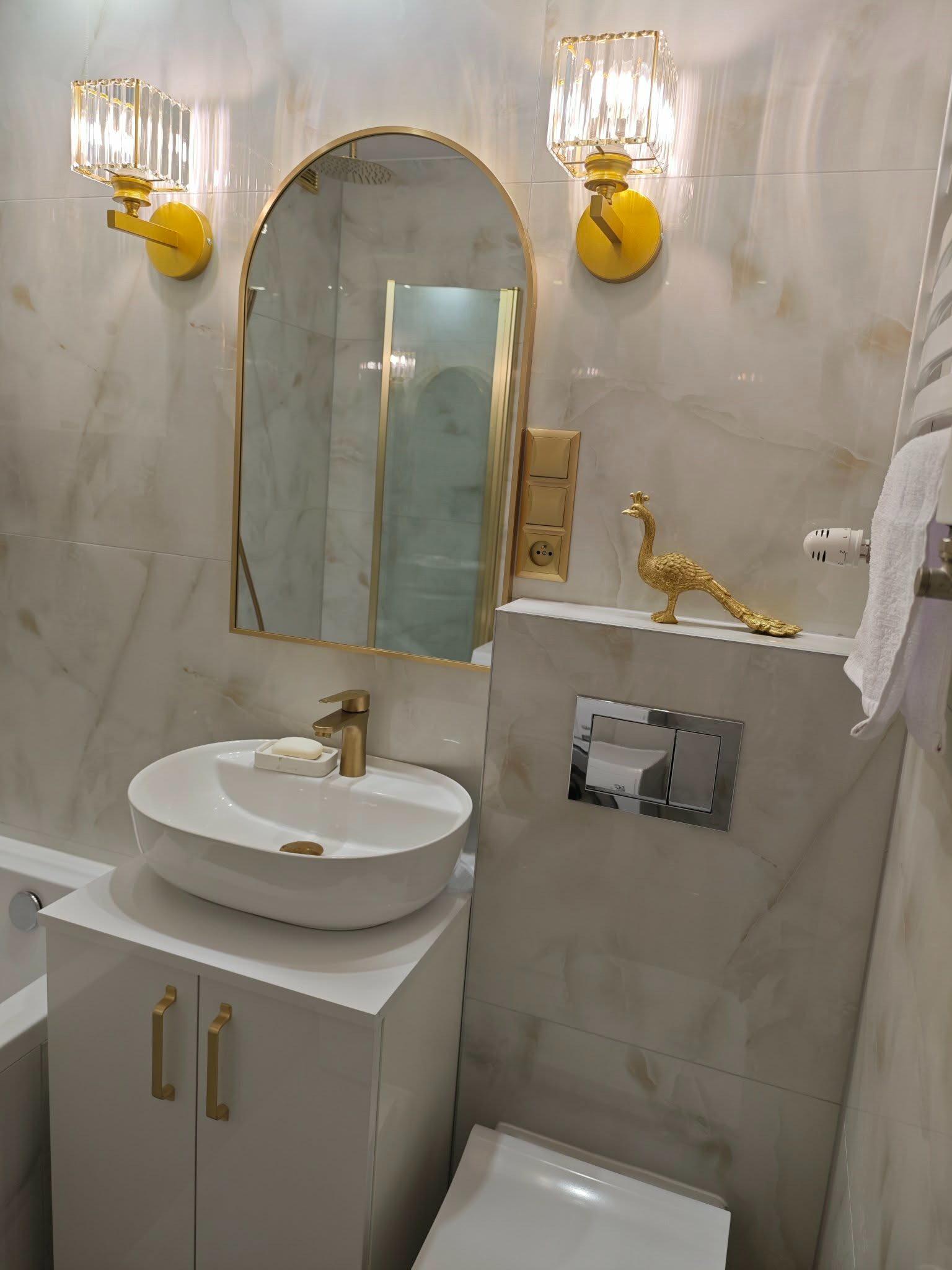 Elegancka łazienka z umywalką nablatową, złotymi akcentami (armatura, oświetlenie, dekoracja pawia) i marmurowymi płytkami. Nowoczesny design z dbałością o detale.