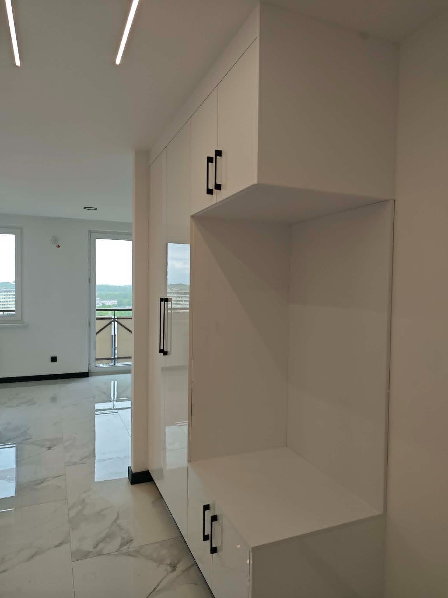Biała zabudowa meblowa z czarnymi uchwytami w przedpokoju, widok z perspektywy podłogi z marmurowym wzorem. Minimalistyczny design, oświetlenie LED na suficie.