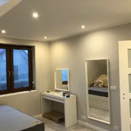 Jasne wnętrze pokoju na poddaszu z szarą ścianą, oknem z ciemną ramą, białymi meblami i lustrami, po remoncie.