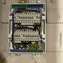 Otwarta skrzynka elektryczna z widocznymi bezpiecznikami Eaton, okablowaniem w różnych kolorach oraz przewodami wychodzącymi ze skrzynki, zamocowana na surowej ścianie z betonu komórkowego.