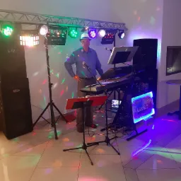 Muzyk w kapeluszu stoi za keyboardem Yamaha, otoczony sprzętem nagłośnieniowym i oświetleniem dyskotekowym, logo zespołu 'Akord' podświetlone neonem.