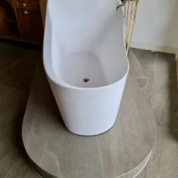 Wolnostojąca, owalna wanna z białego akrylu na podwyższeniu z szarej płyty ceramicznej, z baterią podłogową w kolorze złotym, umywalka z szafką w tle.