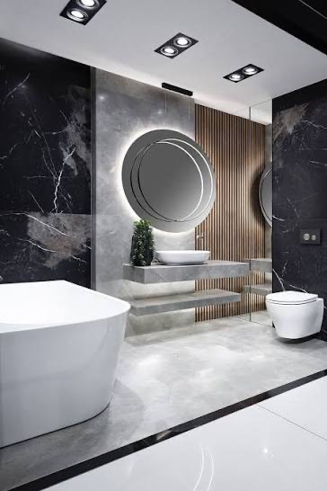 Elegancka łazienka z marmurowymi ścianami, okrągłym lustrem z podświetleniem LED, wolnostojącą wanną i podwieszaną toaletą. Minimalistyczny design z akcentami drewna.