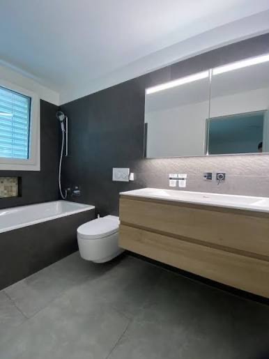 Nowoczesna łazienka z szarymi płytkami, drewnianą szafką pod umywalką i oświetleniem LED nad lustrem. Minimalistyczny design, widoczna wanna i toaleta.