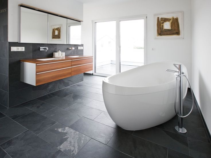 Nowoczesna łazienka z wolnostojącą wanną, ciemną kamienną posadzką i drewnianą szafką pod umywalką. Minimalistyczny design, jasne wnętrze z dużym oknem.