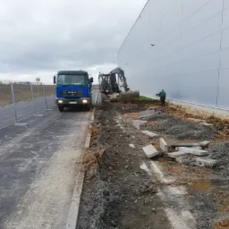 Niebieska ciężarówka MAN i koparka podczas prac przygotowawczych do budowy przy hali magazynowej, widoczne wykopane fragmenty ziemi i ułożone obrzeża chodnika.