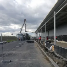 Betoniarka z wysięgnikiem wylewa beton na przygotowany fundament pod obiekt przemysłowy, widoczne zabezpieczenia i pracownicy w czerwonych kombinezonach.