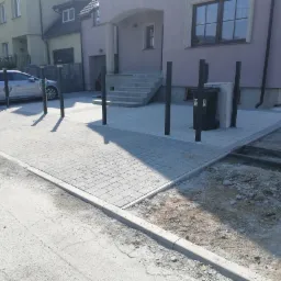 Świeżo ułożona, szara kostka brukowa przed domem jednorodzinnym z czarnymi słupkami, widoczne pozostałości po pracach ziemnych przy krawężniku.