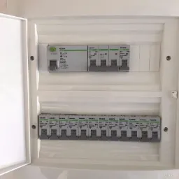 Otwarta biała skrzynka rozdzielcza z zamontowanymi bezpiecznikami Noark Ex9BN B16 i Ex9L-N 40A, widoczne śruby mocujące i fragment ściany.