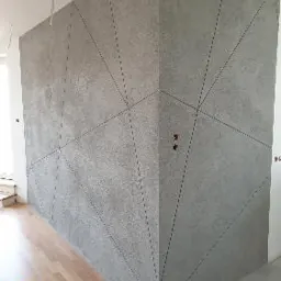 Szara ściana w rogu pomieszczenia z dekoracyjną fakturą i geometrycznym wzorem z cienkich, przerywanych linii. Widoczne gniazdka elektryczne i fragment drewnianej podłogi. W lewym dolnym rogu logo...