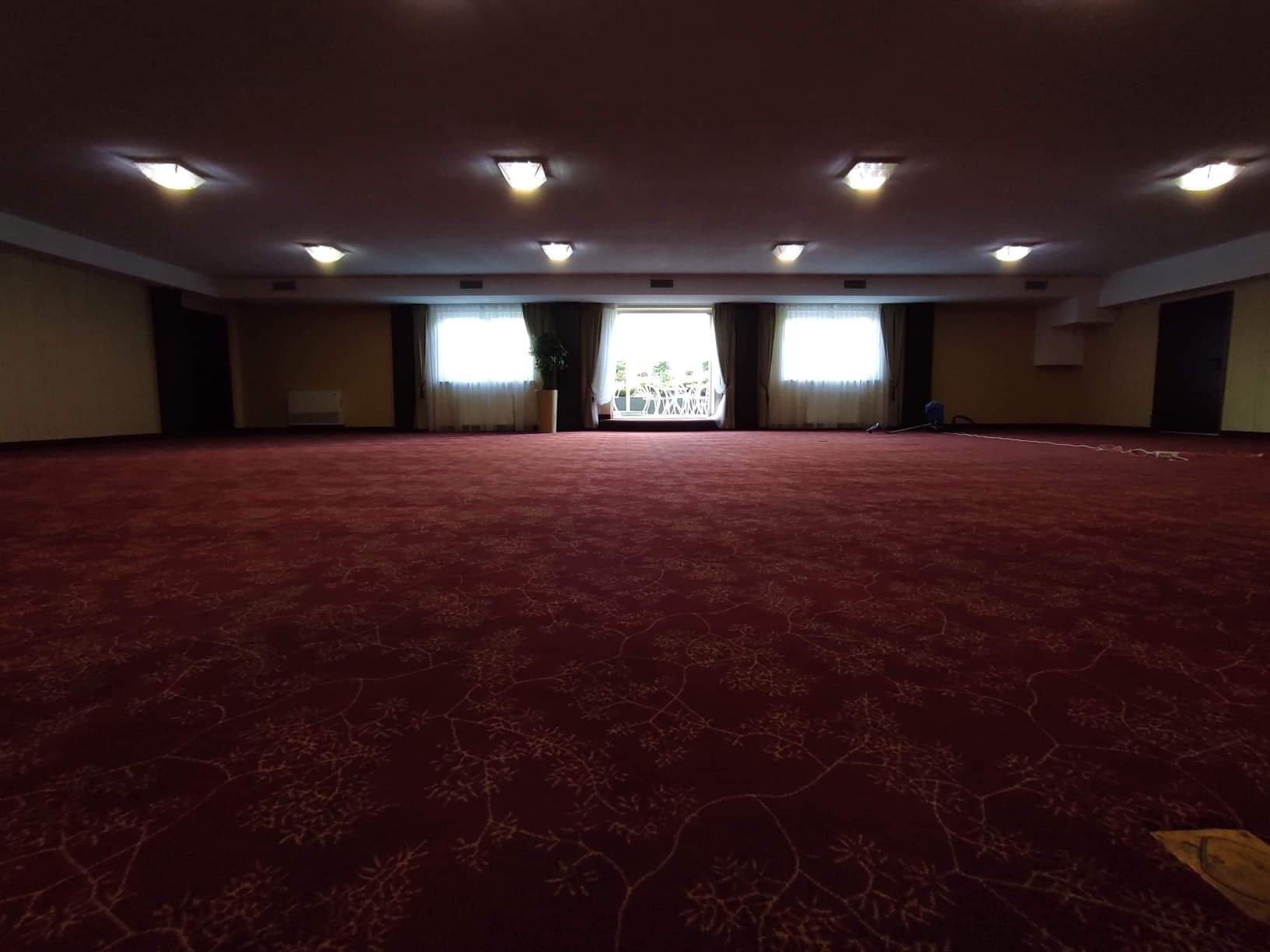 Przestronna sala z czerwonym dywanem w kwiatowy wzór, oświetlona licznymi lampami sufitowymi, z widocznym odkurzaczem na tle okna.