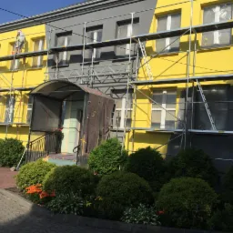 Dwukondygnacyjny budynek z rusztowaniem podczas odnawiania elewacji, część ściany pomalowana na ż&oacute;łto, część na szaro, okna zabezpieczone folią.