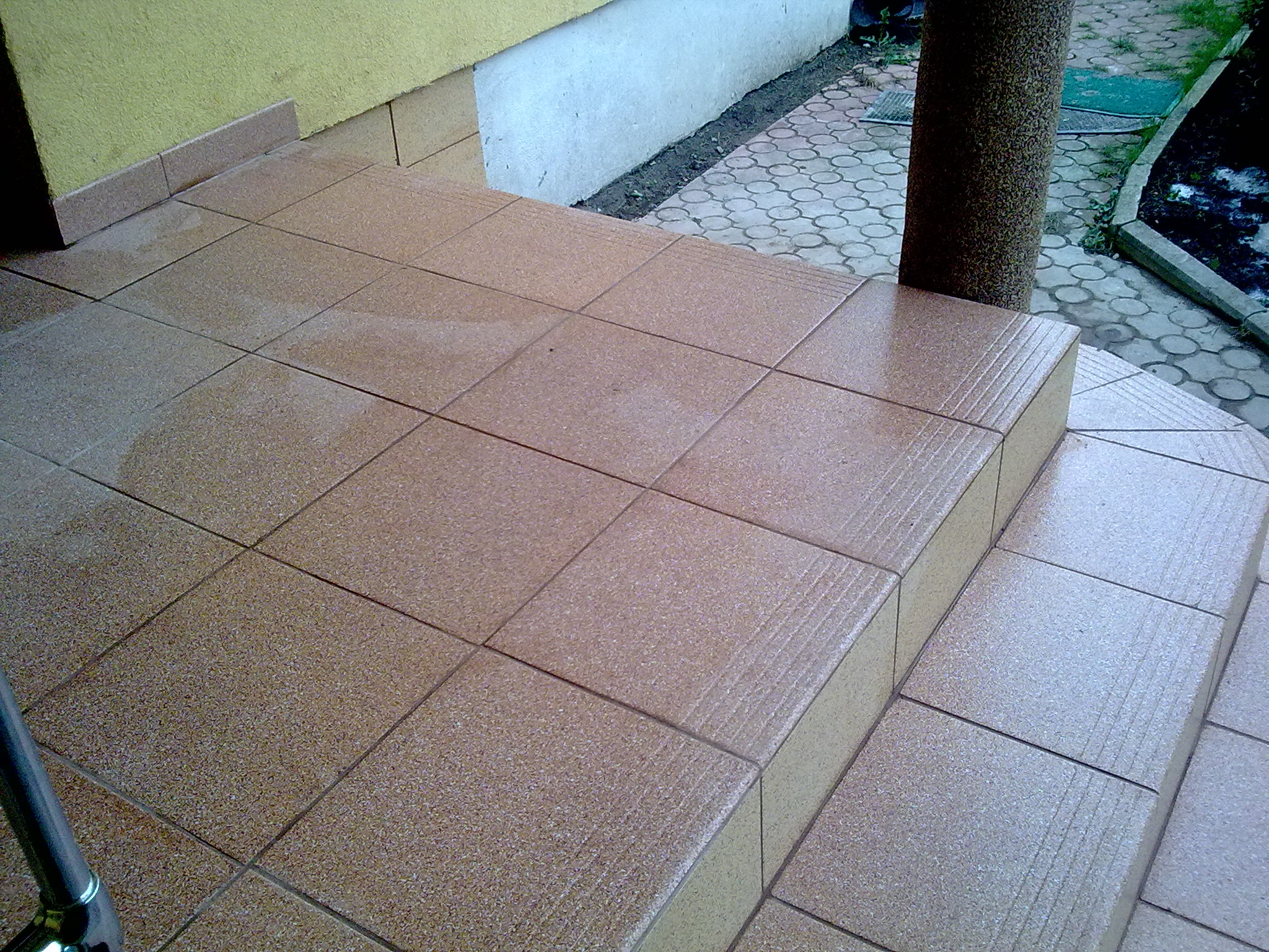 Wyłożone brązowymi płytkami schody wejściowe do domu z kolumną, widoczne płytki chodnikowe na zewnątrz.