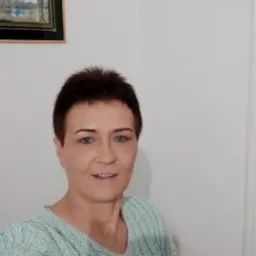 Portret kobiety z krótkimi, ciemnymi włosami i niebieskimi oczami, ubrana w turkusowy sweter z warkoczowym wzorem, na tle białej ściany z obrazem w czarnej ramie