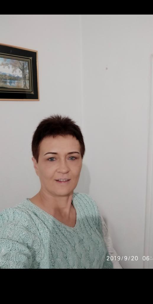 Portret kobiety z krótkimi, ciemnymi włosami i niebieskimi oczami, ubrana w turkusowy sweter z warkoczowym wzorem, na tle białej ściany z obrazem w czarnej ramie