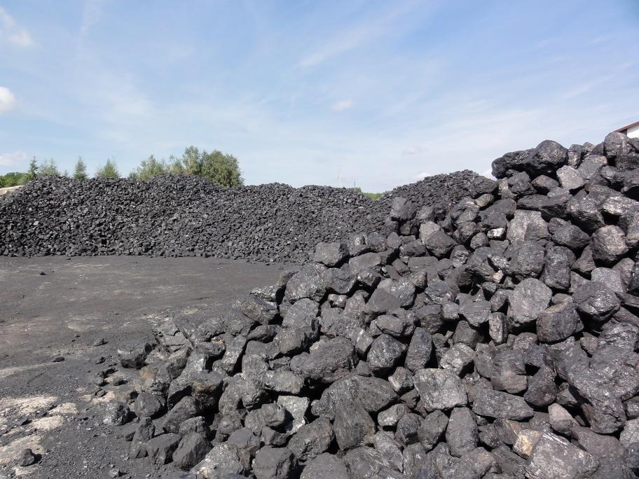Dwa duże pryzmy węgla kamiennego na placu składowym, z widocznym pyłem węglowym na podłożu i zielenią w tle.
