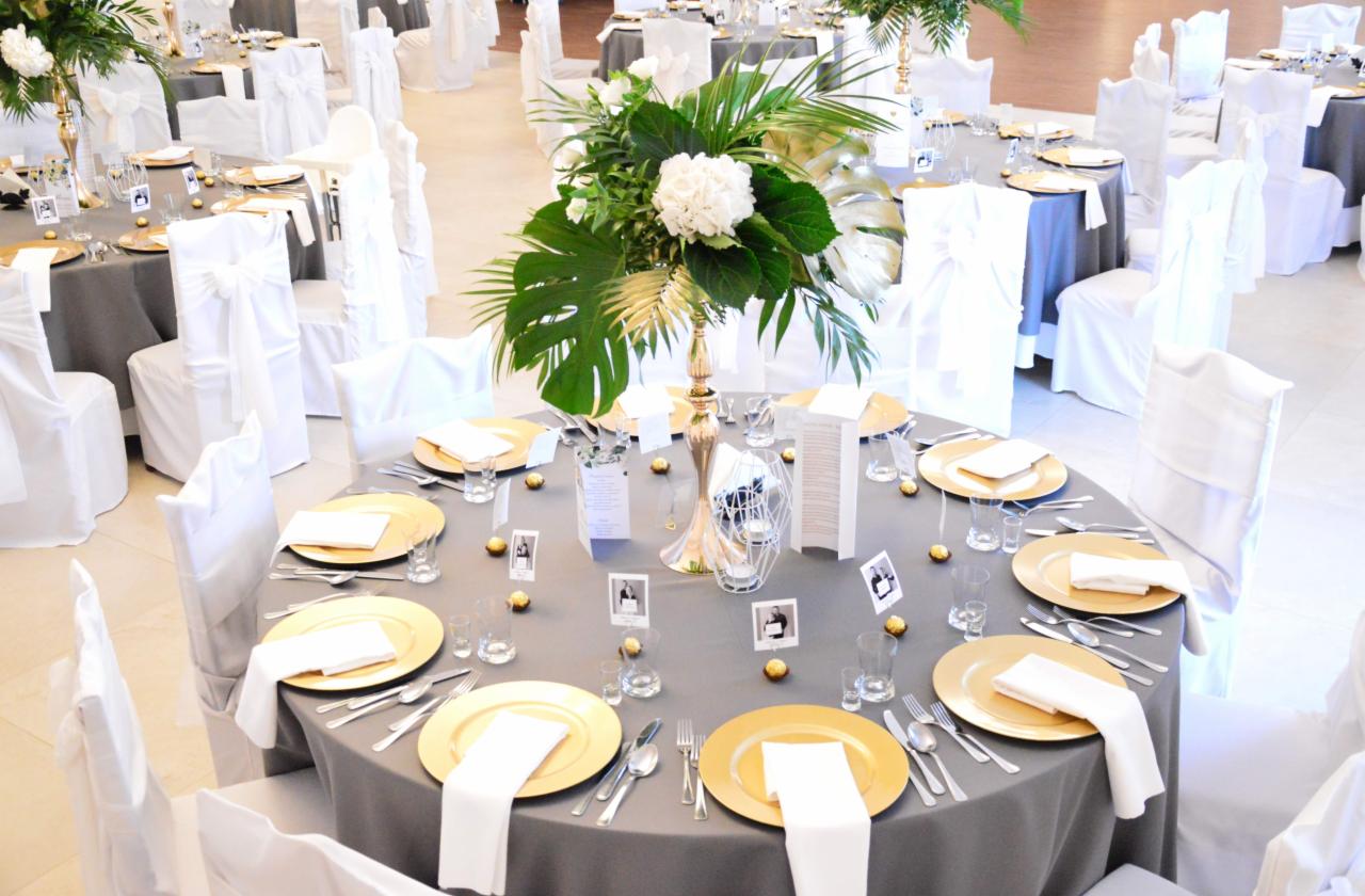 Elegancko nakryty stół z szarą obrusą, złotymi talerzami, białymi serwetkami, szklankami, sztućcami i dekoracjami kwiatowymi w wysokim złotym wazonie, dodatkowo czekoladki i zdjęcia w ramkach.