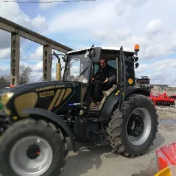 Czarny ciągnik Farmtrac 9120DTn z otwartymi drzwiami kabiny, w której siedzi mężczyzna. W tle widoczne budynki i sprzęt rolniczy.