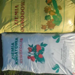 Dwa worki ziemi: żółty worek z ziemią ogrodową i biały z ziemią kwiatową uniwersalną, leżące na trawie.