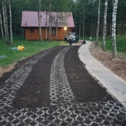 Budowa podjazdu z ażurowych płyt betonowych wypełnianych ziemią, widoczny fragment chodnika z jasnej kostki, w tle drewniany domek letniskowy i brzozowy lasek.