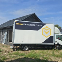 Okna Drzwi Rolety itp. Krzysztof Brzychcy - Ciężarówka z logo firmy oferującej okna, drzwi i rolety zaparkowana przed domem w trakcie montażu okien. Na pierwszym planie widoczne okna z białymi ramami opierające się o ścianę budynku.