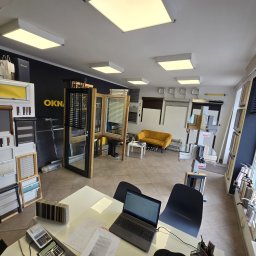 Okna Drzwi Rolety itp. Krzysztof Brzychcy - Wnętrze salonu z ekspozycją okien i drzwi, widoczny laptop na biurku, próbki kolorów i materiałów, oraz kanapa dla klientów w tle.