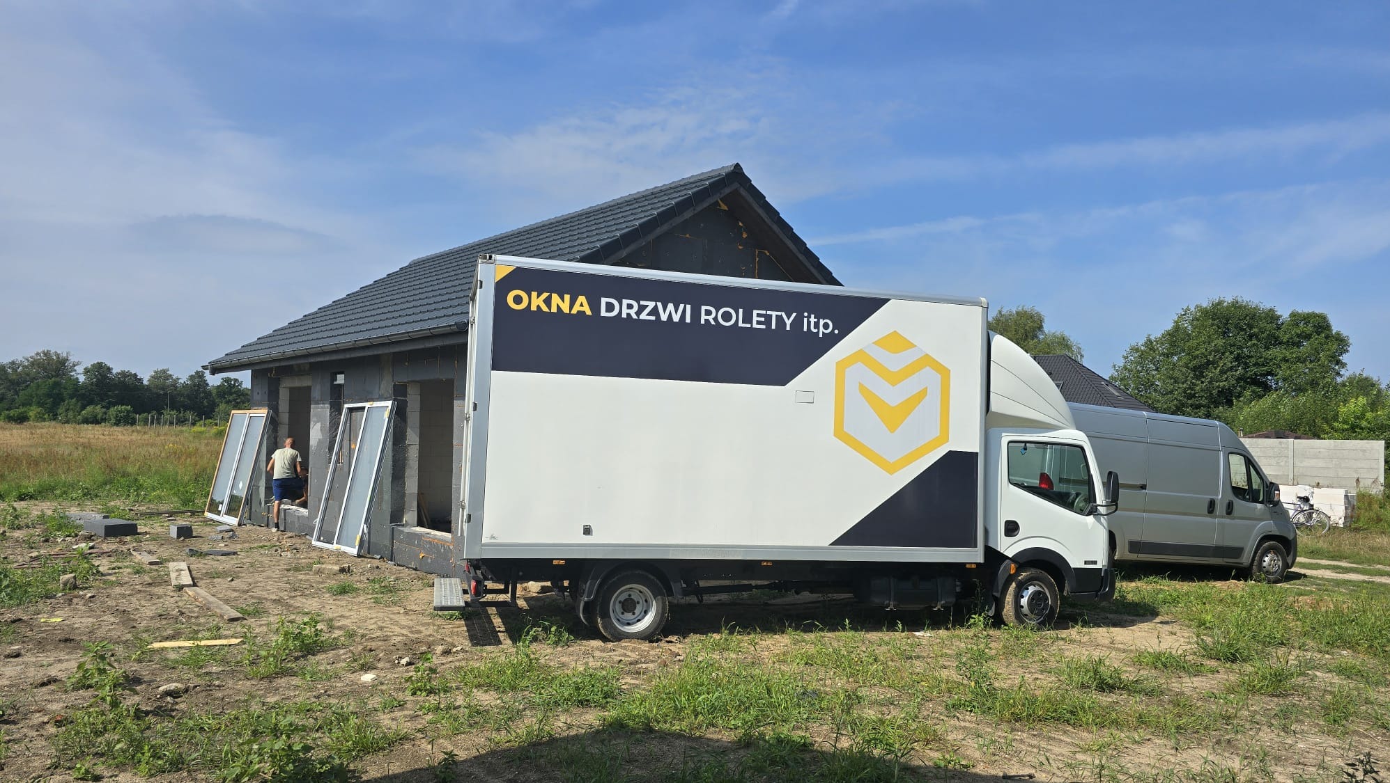 Ciężarówka z logo firmy oferującej okna, drzwi i rolety zaparkowana przed domem w trakcie montażu okien. Na pierwszym planie widoczne okna z białymi ramami opierające się o ścianę budynku.