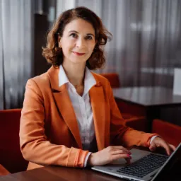 Kobieta w pomarańczowej marynarce, siedząca przy ciemnym stole i pracująca na laptopie. Uśmiechnięta, z krótkimi kręconymi włosami. W tle zasłony i stolik.