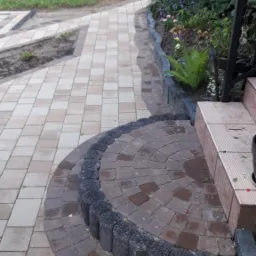 Kaskadowa ścieżka z kostki brukowej w odcieniach beżu i brązu, obrzeża z ciemnych, okrągłych elementów, prowadząca do schodów i ogrodu z roślinami.