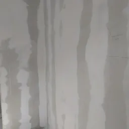 Niedokończona ściana narożna z płyt kartonowo-gipsowych, z widocznymi śladami szpachlowania i odręcznym napisem w prawym górnym rogu.