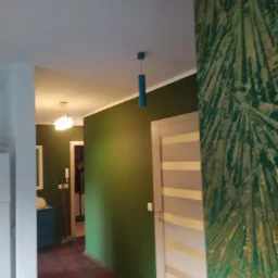 Wnętrze mieszkania z zielonymi ścianami, drzwi z poziomymi panelami i fragment ściany z dekoracyjnym motywem liści palmowych w odcieniach zieleni i złota. W głębi widoczny korytarz z oświetleniem.