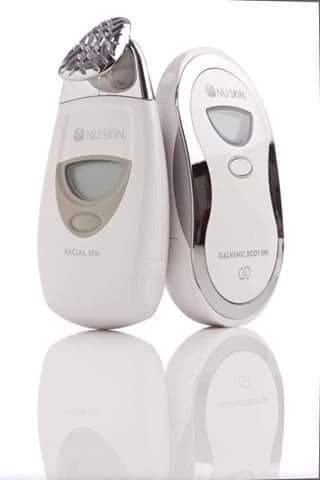 Dwa białe urządzenia kosmetyczne marki Nu Skin: Facial Spa z metalową końcówką i wyświetlaczem oraz Galvanic Body Spa, oba z srebrnymi akcentami, ustawione obok siebie na błyszczącej powierzchni...